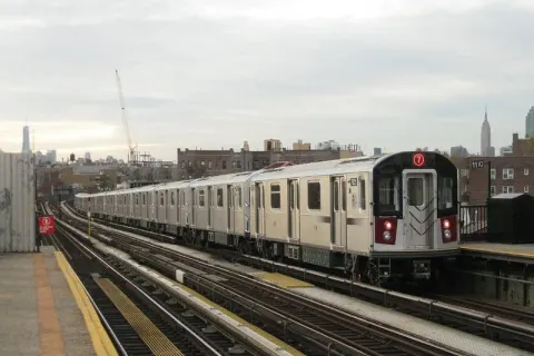 A New York City Transit "7" subway train arriving at an elevated station, with the Manhattan skyline in the background.